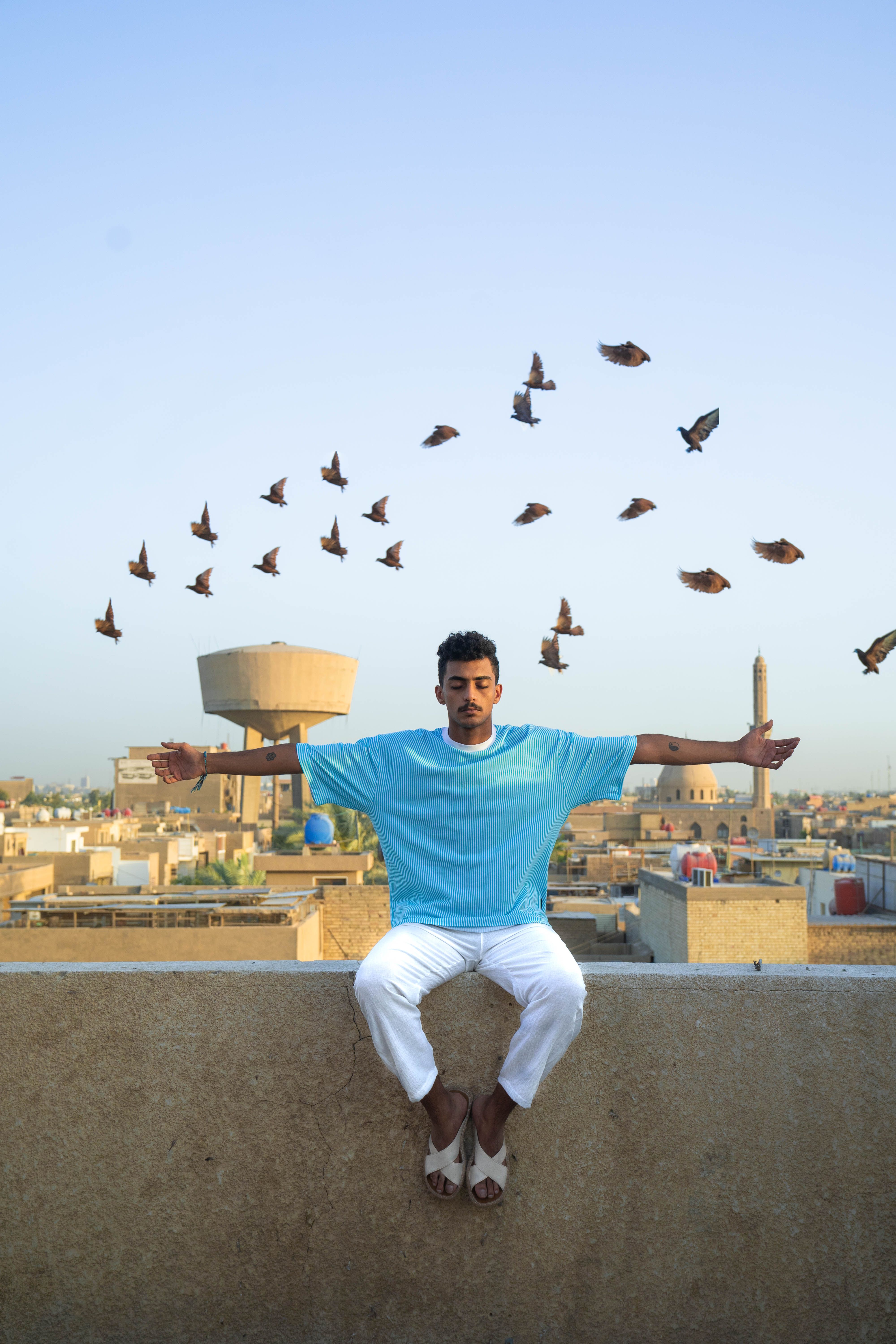 A young man sits on a rooftop with arms outstretched as birds fly behind him.
