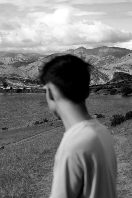 Fotomural Una foto en escala de grises que muestra a un hombre contemplando un tranquilo lago hacia montañas distantes.