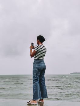 Woman in denim jeans and striped shirt taking a photo of the ocean under a cloudy sky.