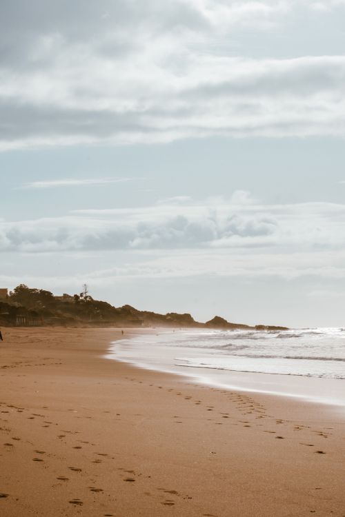 Free Serene sandy beach with footprints and soft waves under a cloudy sky. Stock Photo