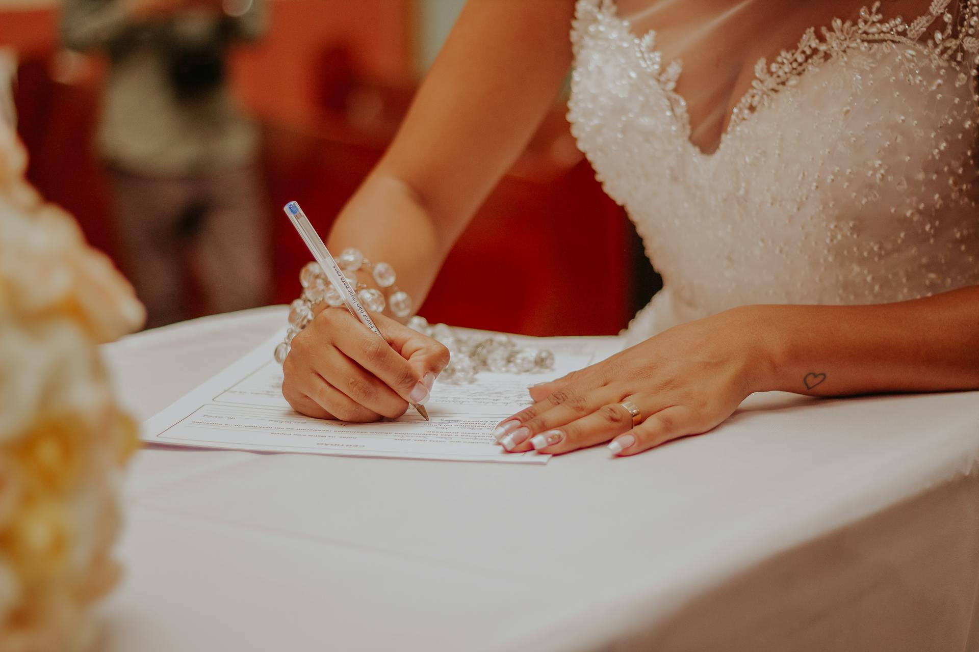 A bride signs a marriage document during her wedding ceremony with intricate jewelry visible.