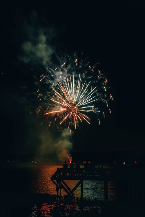 Free Colorful fireworks burst over a lake with silhouetted people watching from a dock at night. Stock Photo