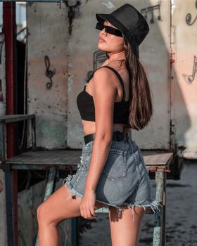 Fashionable woman wearing denim shorts and black hat poses outdoors in sunlight. Summer style.