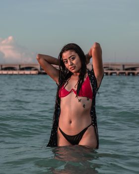 A confident woman in a red bikini stands in the ocean water at sunset.