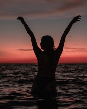 Silhouette of a woman in bikini with arms raised, posing in the sea at sunset.