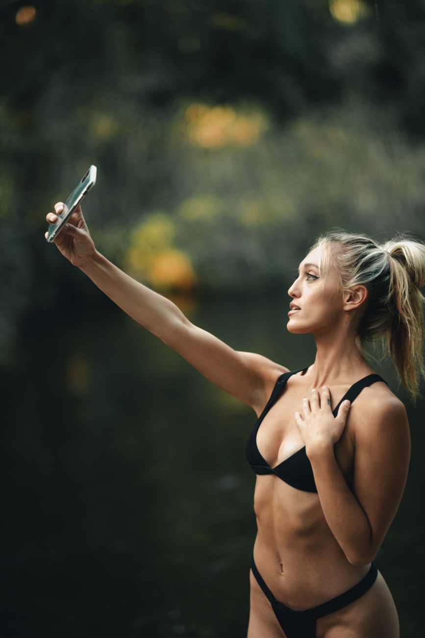 Blonde woman in a bikini taking a selfie by a serene outdoor setting.