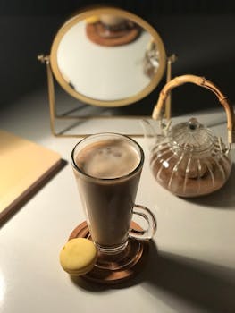 A sophisticated coffee setup with a glass mug, yellow macaron, teapot, and a round mirror reflection.