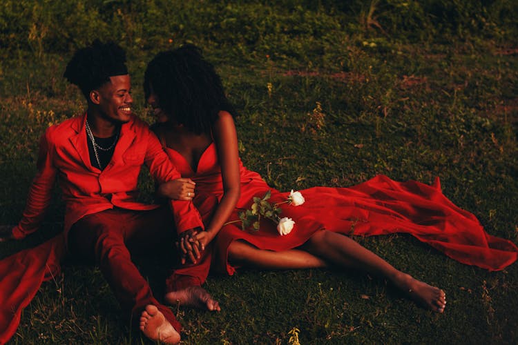 Smiling Couple In Red Clothing Sitting On Grass And Hugging