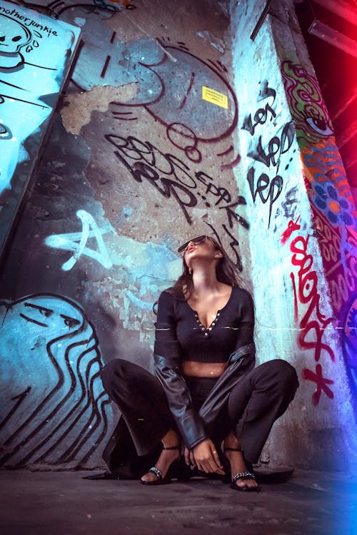 Woman in Black Long Sleeves and Black Pants Sitting Beside a Graffiti Wall