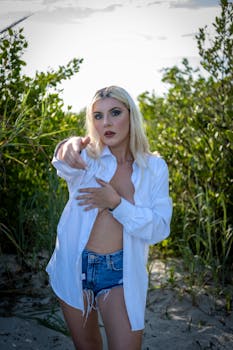 A blonde woman in a white shirt and denim shorts poses confidently in a natural setting.