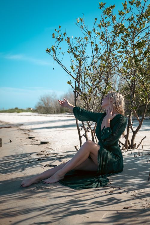 Free A woman in a green dress sits on a sunny sandy beach surrounded by plants. Stock Photo