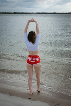 Woman in lifeguard shorts stretching on a beach, facing the sea under a cloudy sky.
