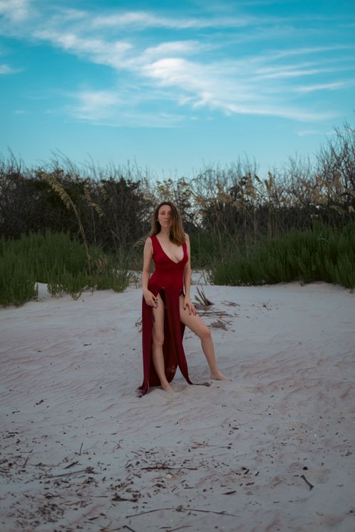 Free A woman poses barefoot in a sleeveless red dress on a sandy beach surrounded by greenery. Stock Photo