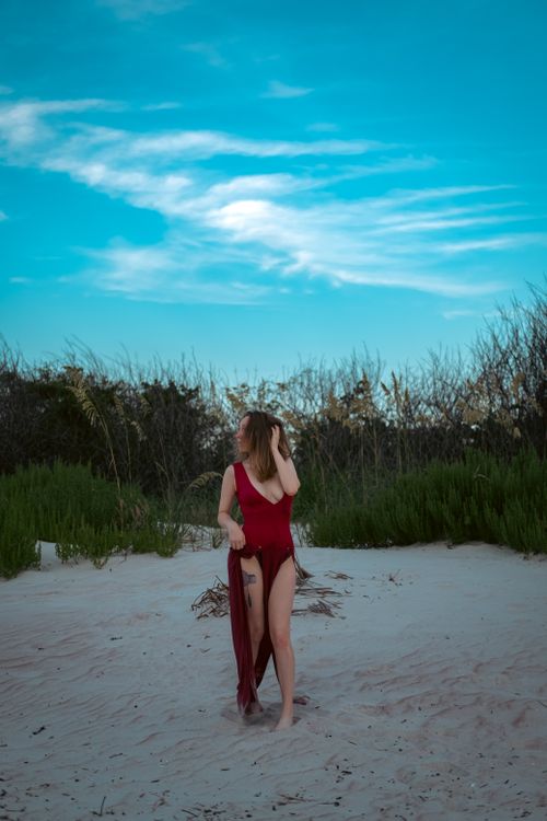 Free A woman in a red dress stands on a sandy beach with lush greenery under a twilight sky. Stock Photo