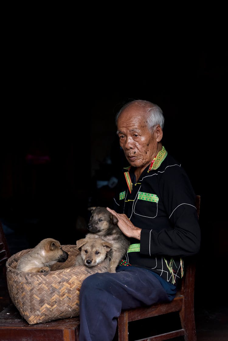 An Elderly Man Sitting On A Wooden Chair While Petting His Dogs