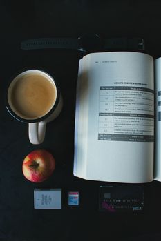 Flat lay featuring coffee, open book, apple, watch, and tech items on a dark surface.