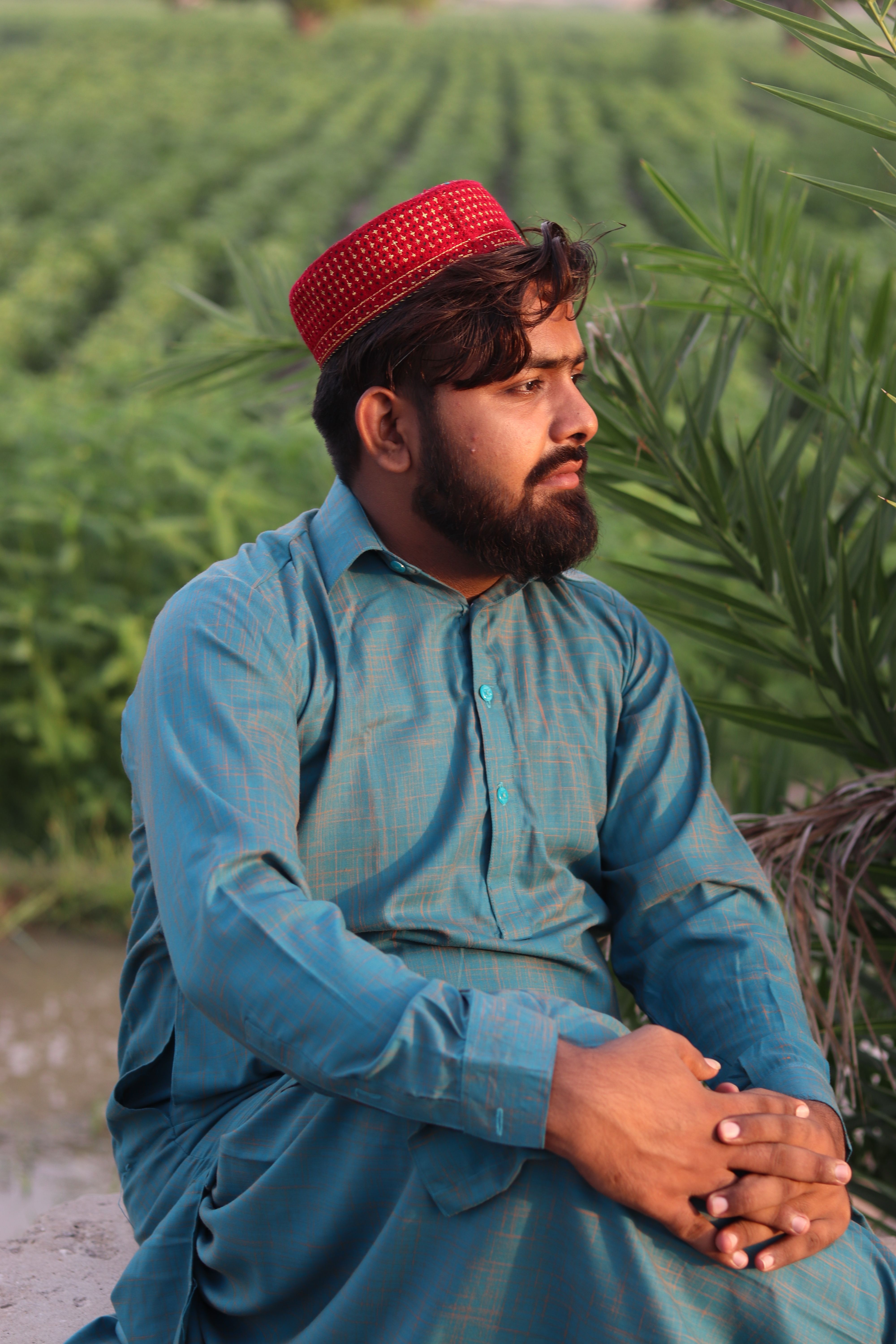 A man with a red cap sits cross-legged in a field, wearing blue traditional attire.