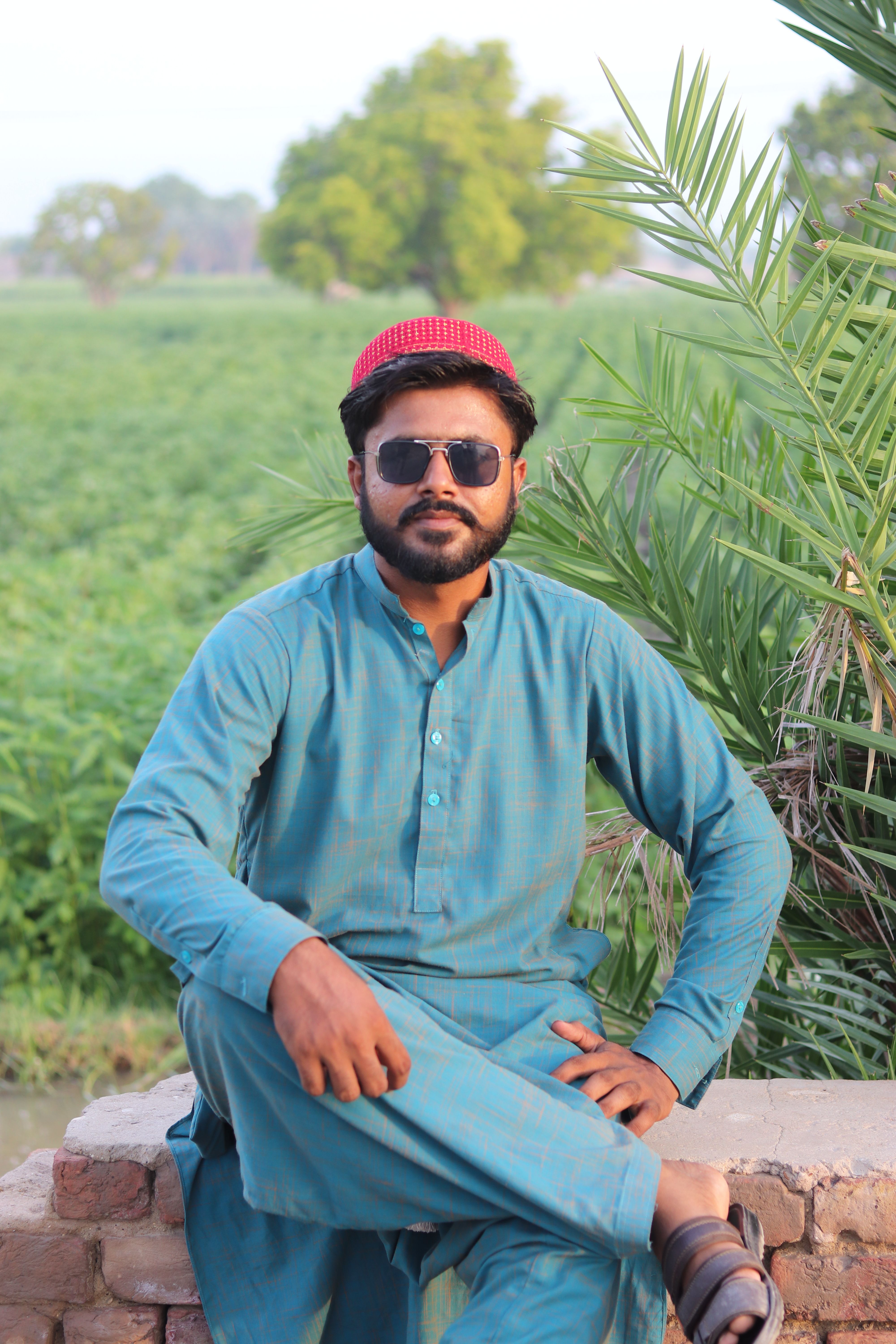 A man in traditional attire sitting outdoors on a sunny day with greenery in the background.