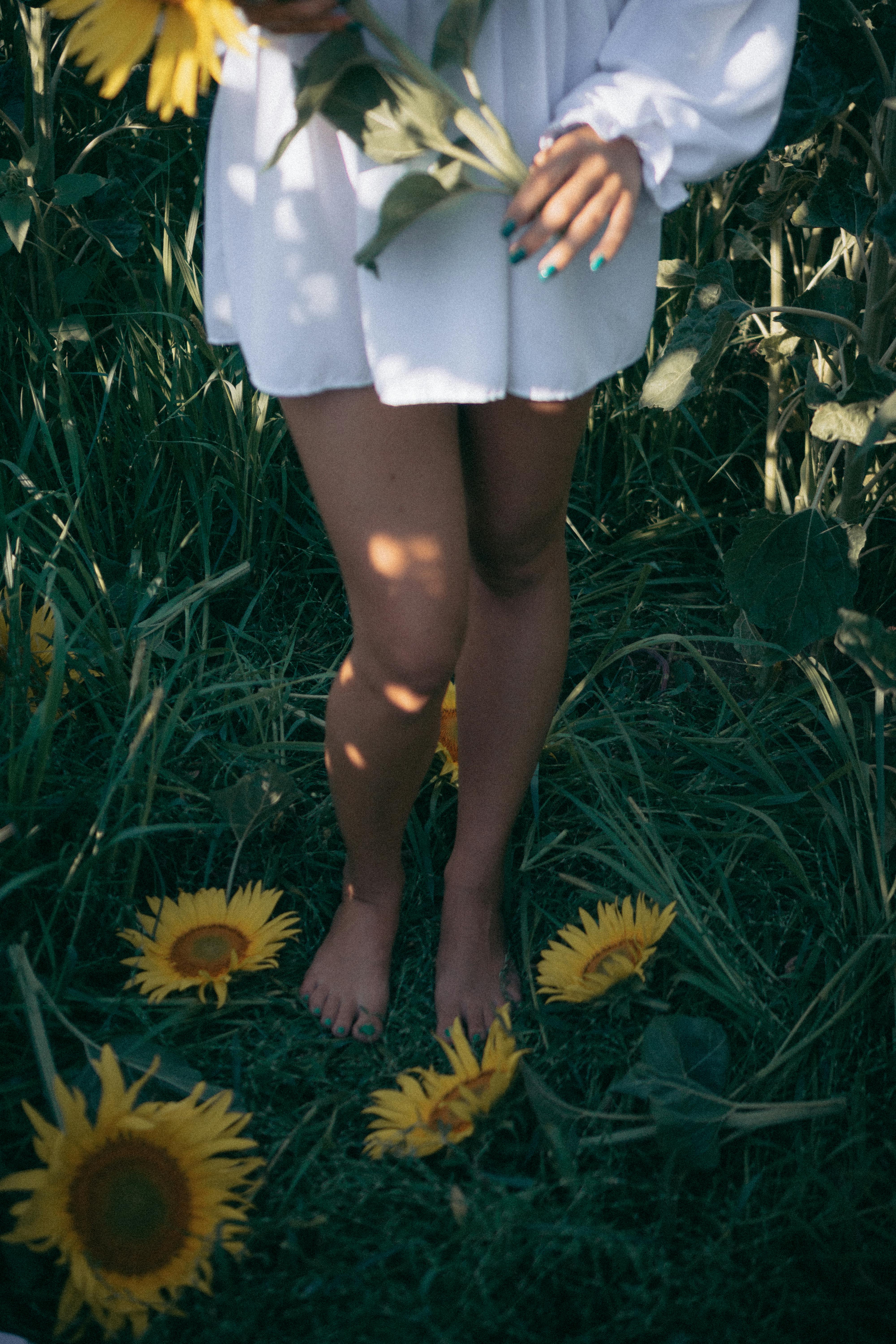 A woman stands barefoot among sunflowers, holding blossoms in a summer field.