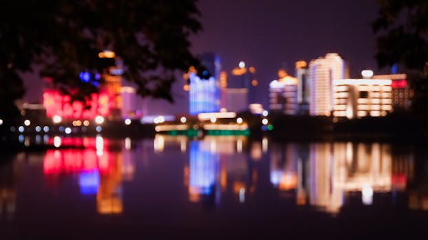 A blurred night cityscape with colorful illuminated buildings reflecting on water.