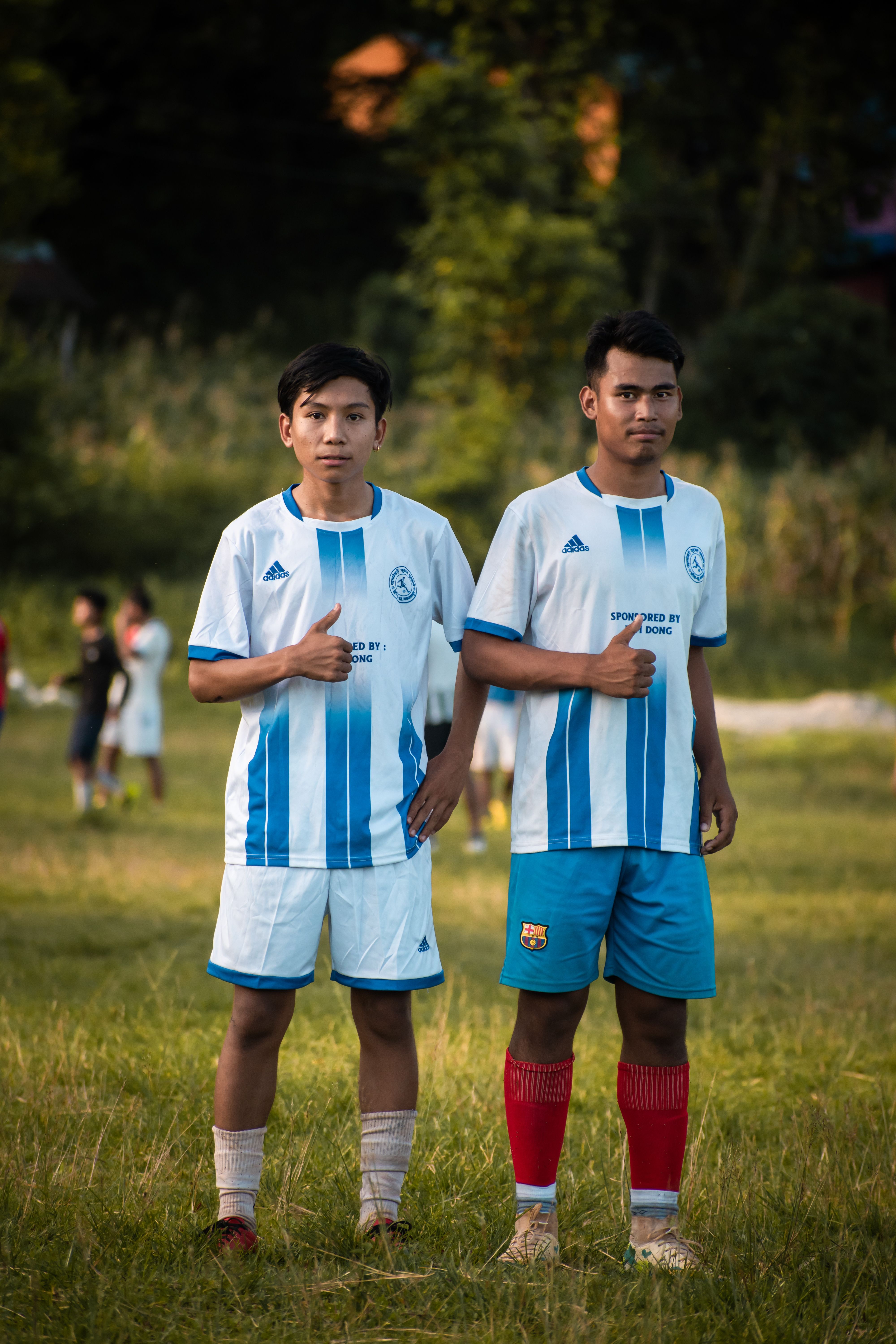 Two young soccer players posing confidently on a grass pitch, thumbs up.