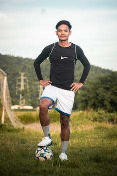 Asian male athlete posing confidently with a soccer ball on a grassy field.