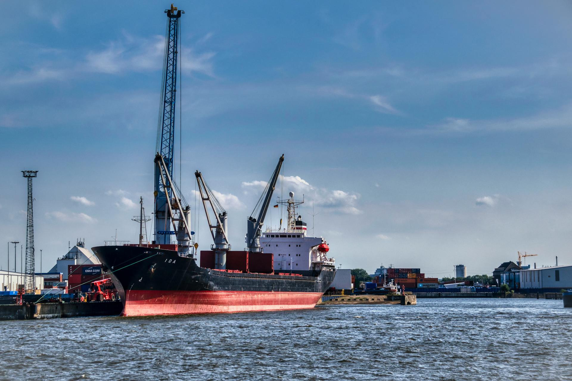 View of a cargo ship docked at a busy port under a clear blue sky, surrounded by cranes and containers.