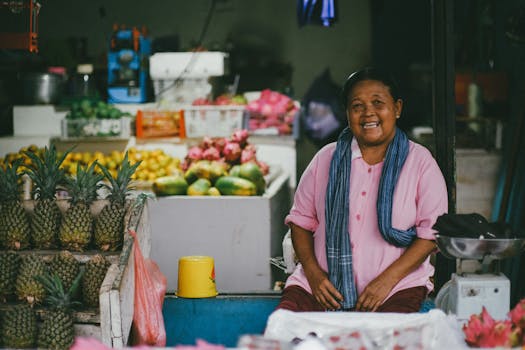 Smiling woman sitting in a colorful fruit market stall with pineapples and other fruits.