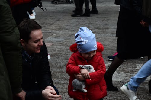 Baby in Red Jacket Holding a Rabbit Beside a Man Sitting in Black Jacket