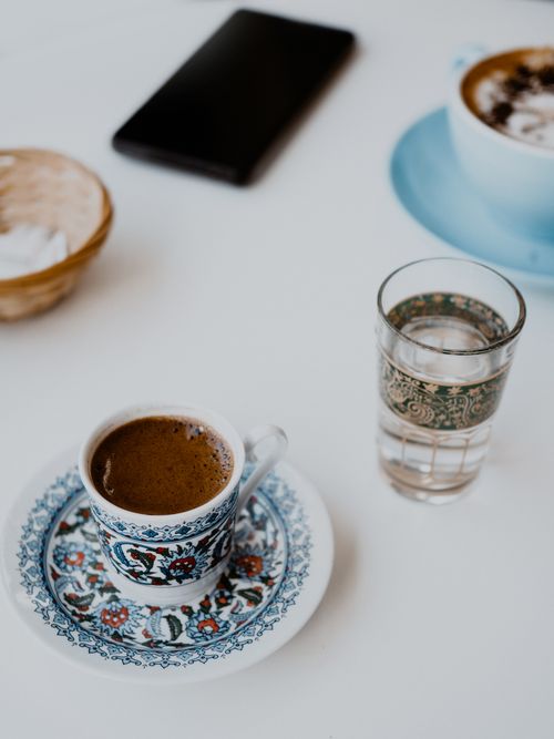Free A beautifully decorated porcelain coffee cup with a matching saucer on a white table, complemented by a glass of water and smartphone. Stock Photo