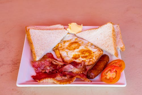Photo of a White Plate with Toasts, Fried Eggs, Fried Becon, Butter, Slices of Tomatoes and a Sausage