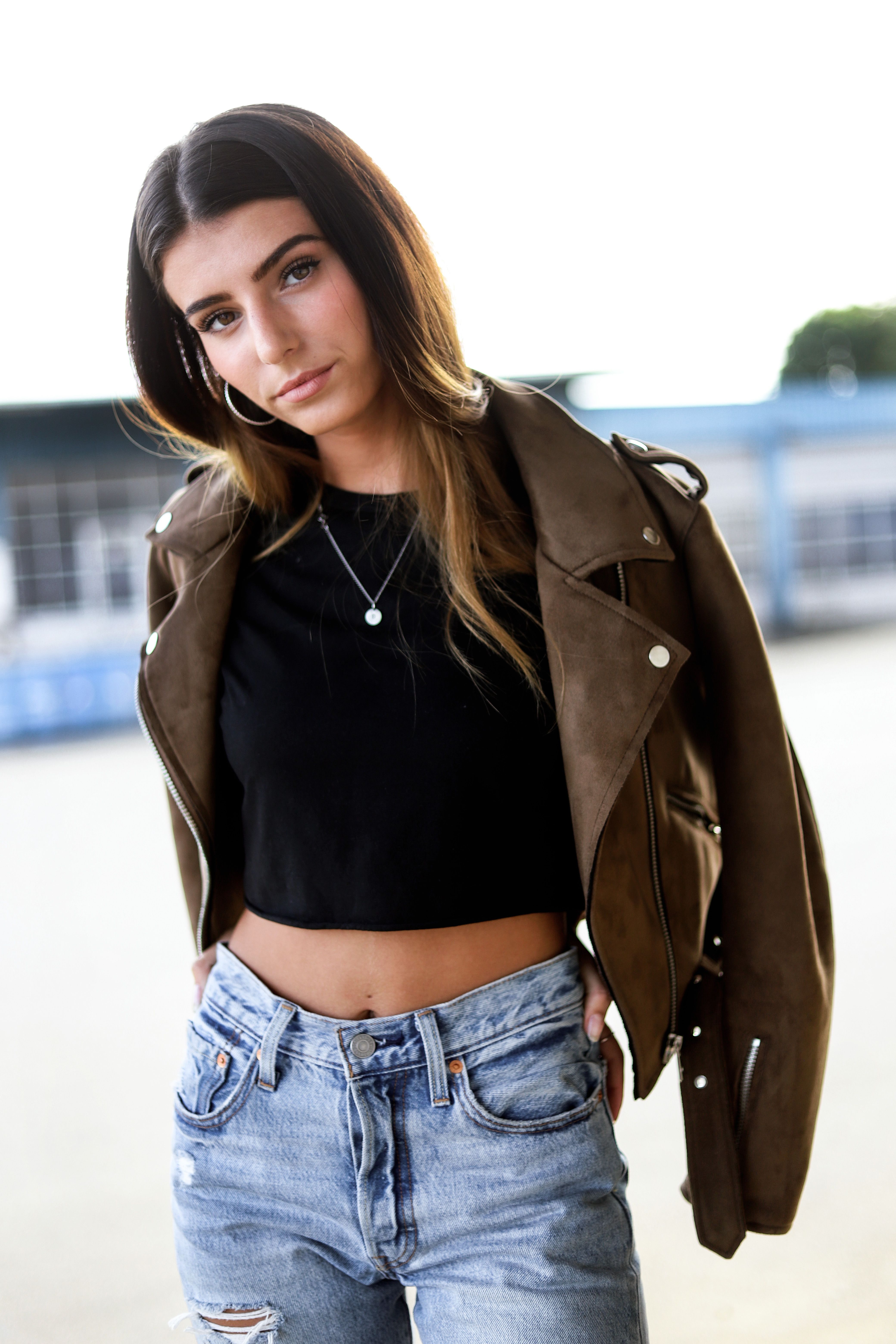 A fashionable woman poses outdoors in a brown jacket and denim, exuding confidence and style.