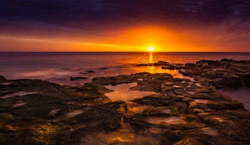 Free Stunning sunset over rocky shore with vibrant orange and purple sky creating a dramatic coastal seascape. Stock Photo