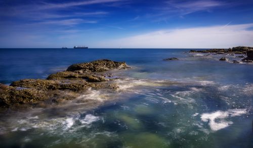 Free Tranquil ocean scene with rocky shores, clear blue skies, and a distant ship on the horizon. Stock Photo