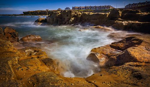 Free Captivating view of coastal rocks and waves with distant townhouses under a clear sky. Stock Photo