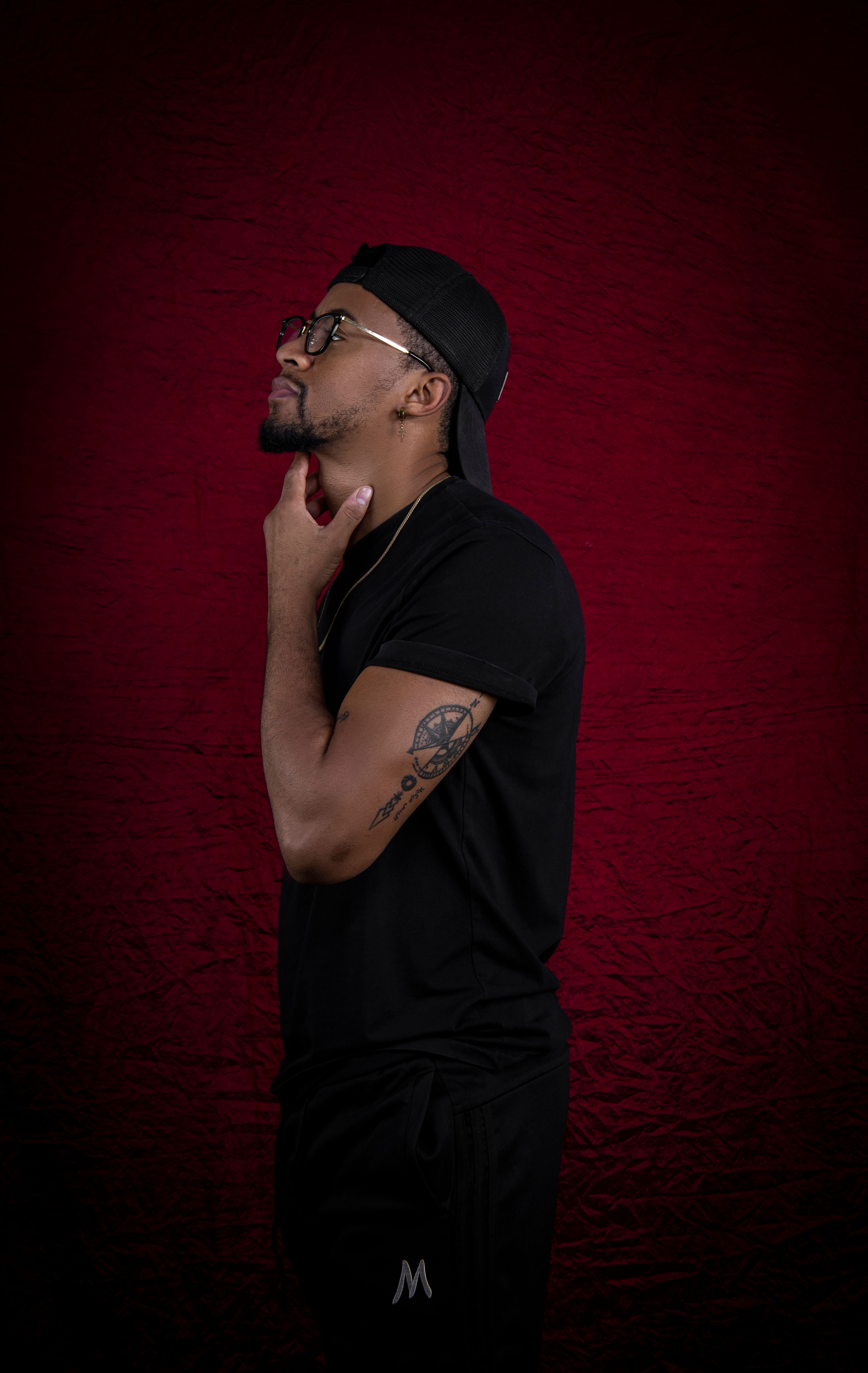 Side profile of a man in a black outfit and cap posing contemplatively in a studio setting.