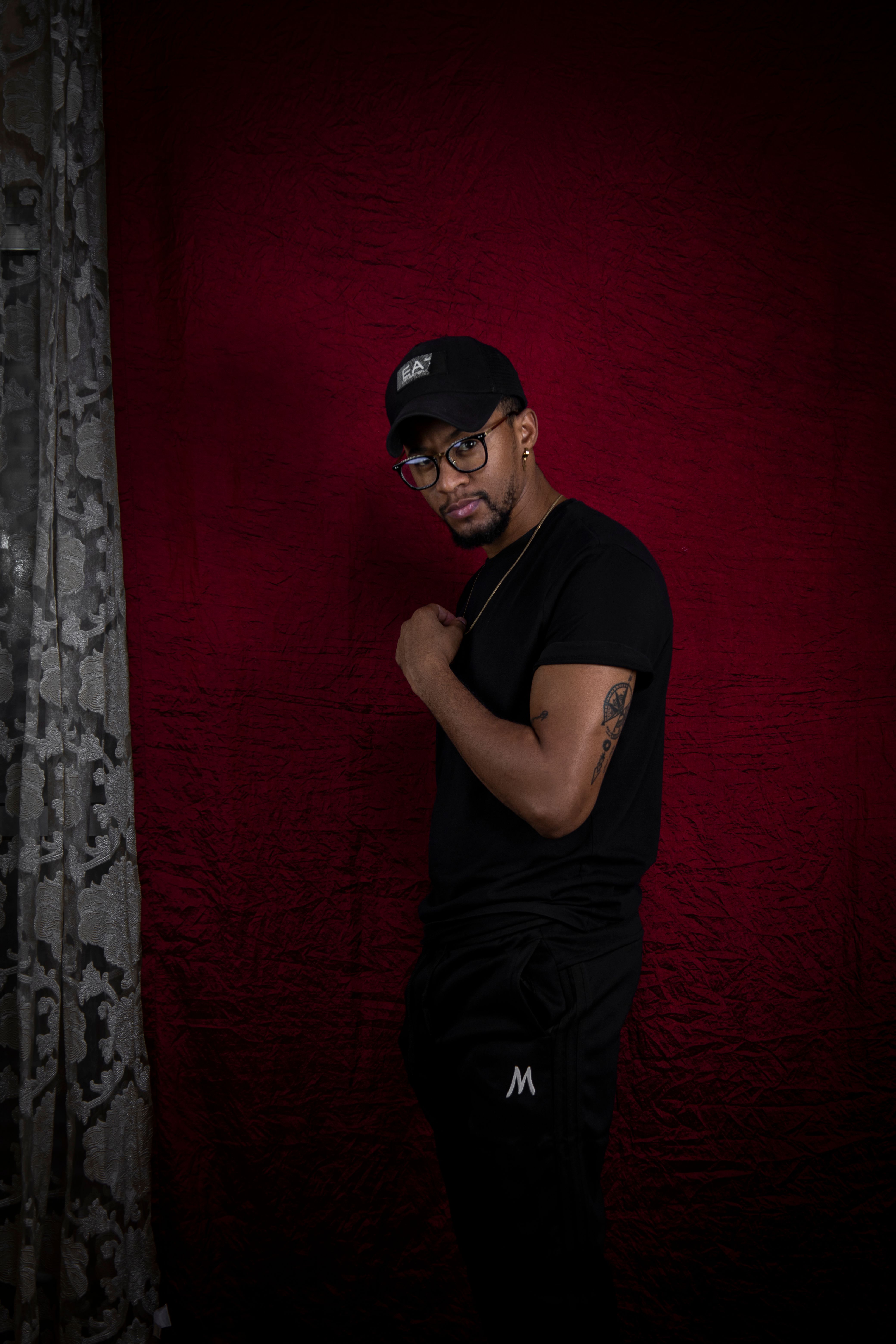 Portrait of a stylish man in a black outfit and cap, standing confidently in a studio setting.