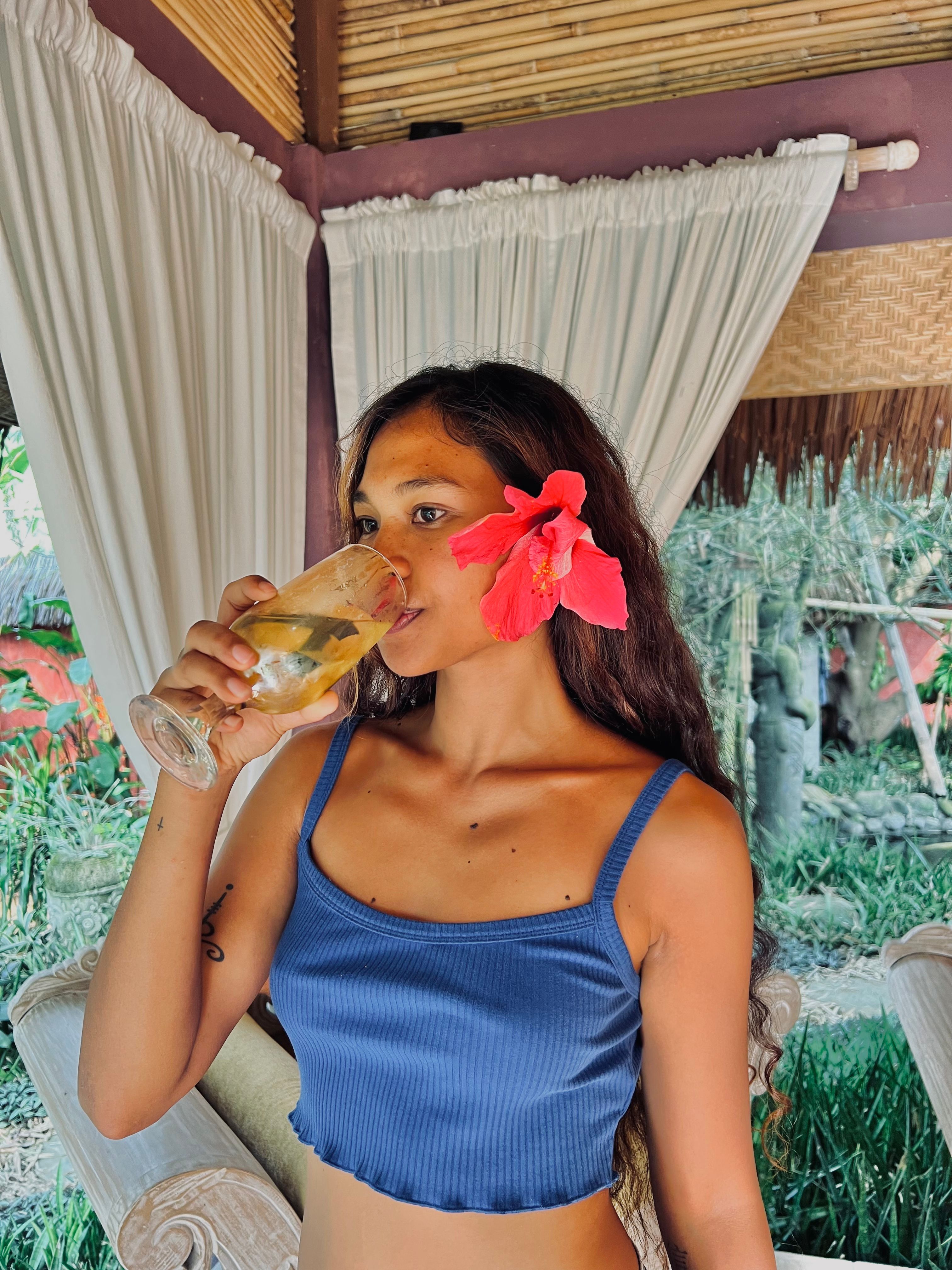A woman in a blue top drinks a beverage, adorned with a hibiscus flower, in an outdoor tropical setting.