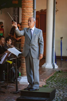 Senior conductor in gray suit leading an orchestra outdoors in Venezuela.