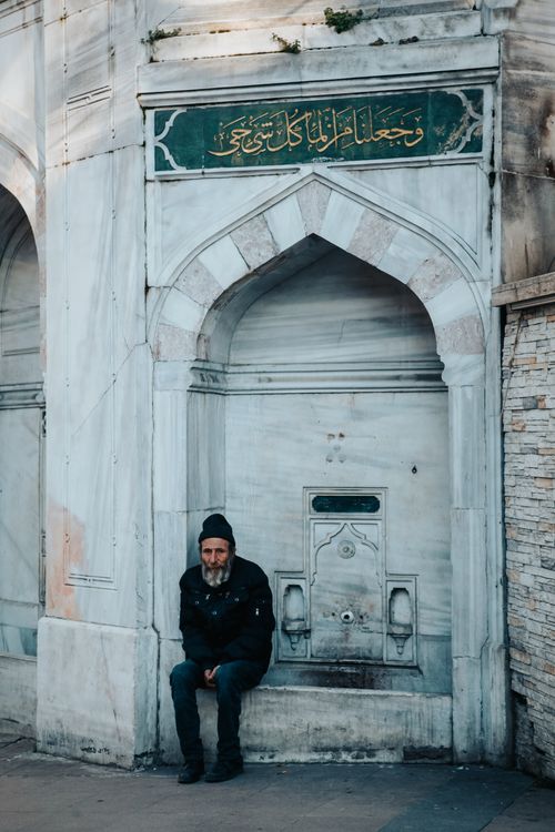 Free An elderly man sits in a black jacket by an ornate stone fountain with Arabic script. Stock Photo