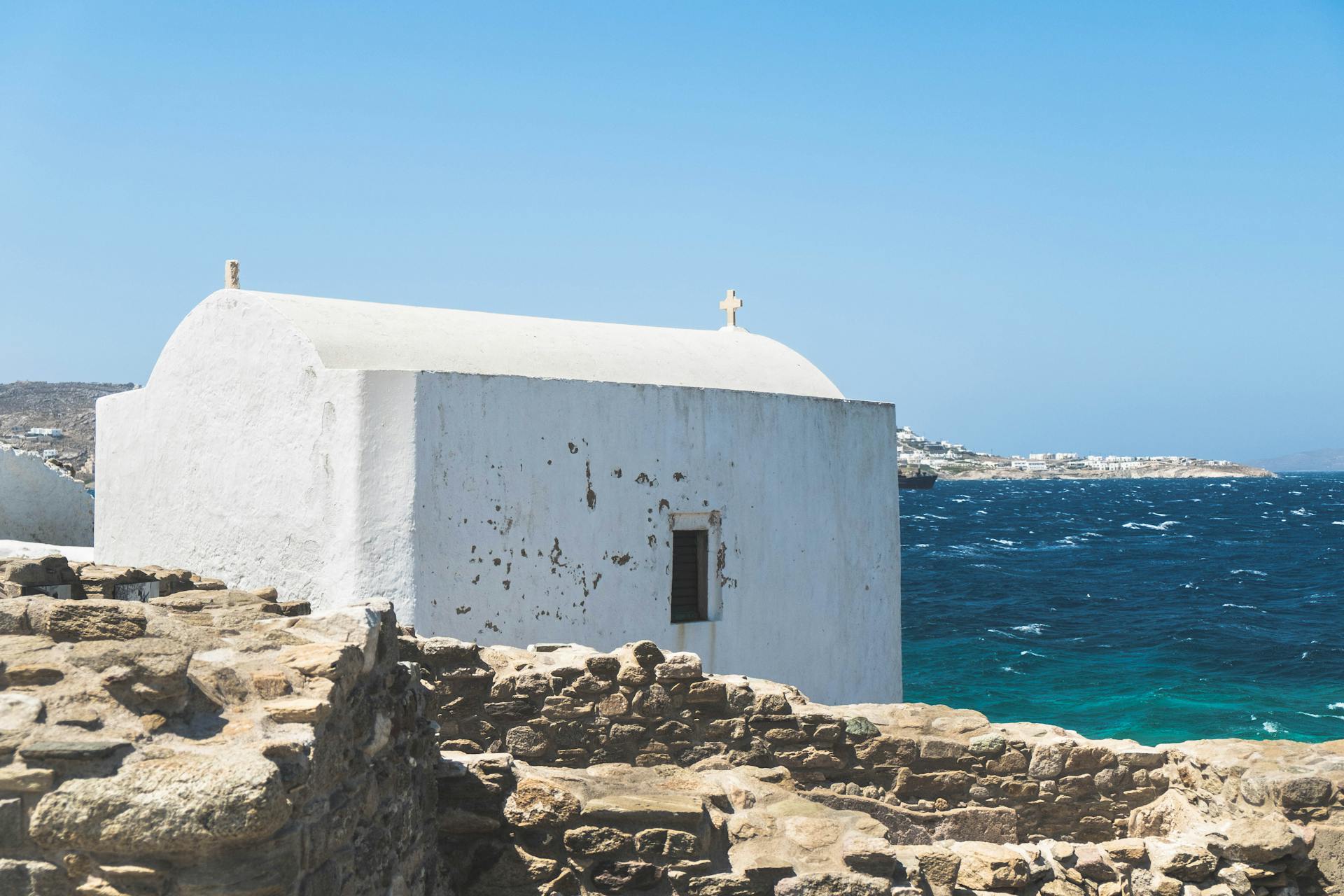 A historic white chapel on Mykonos coastline with vibrant blue Aegean Sea waters.