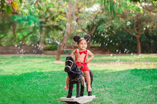 A joyful child with mouse ears sits on a toy horse surrounded by bubbles in a sunny park.