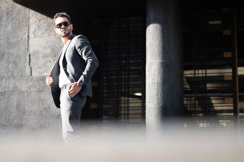 Free A stylish man in sunglasses and gray suit posing confidently outdoors with a modern, urban backdrop. Stock Photo