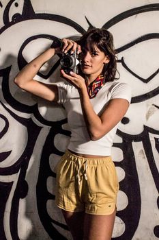 Woman in white shirt and shorts posing with a camera against graffiti art.