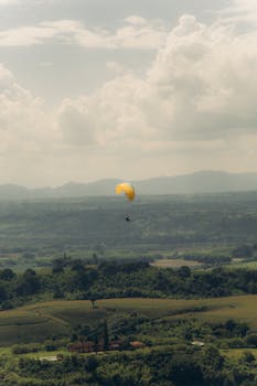 A paraglider with a yellow chute soaring over lush green fields under cloudy skies.