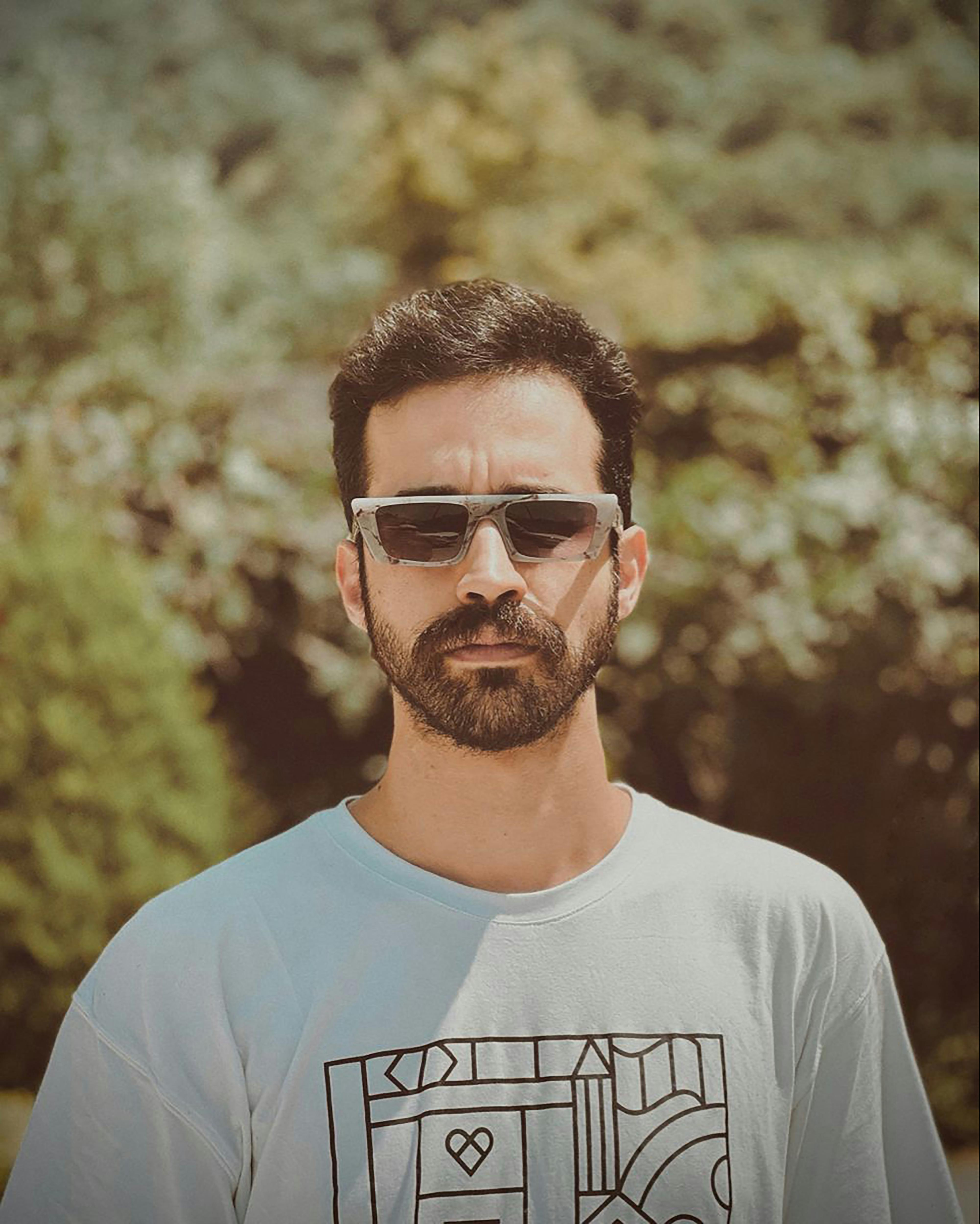 A stylish man wearing sunglasses and a graphic white shirt stands confidently outdoors with blurred trees in the background.