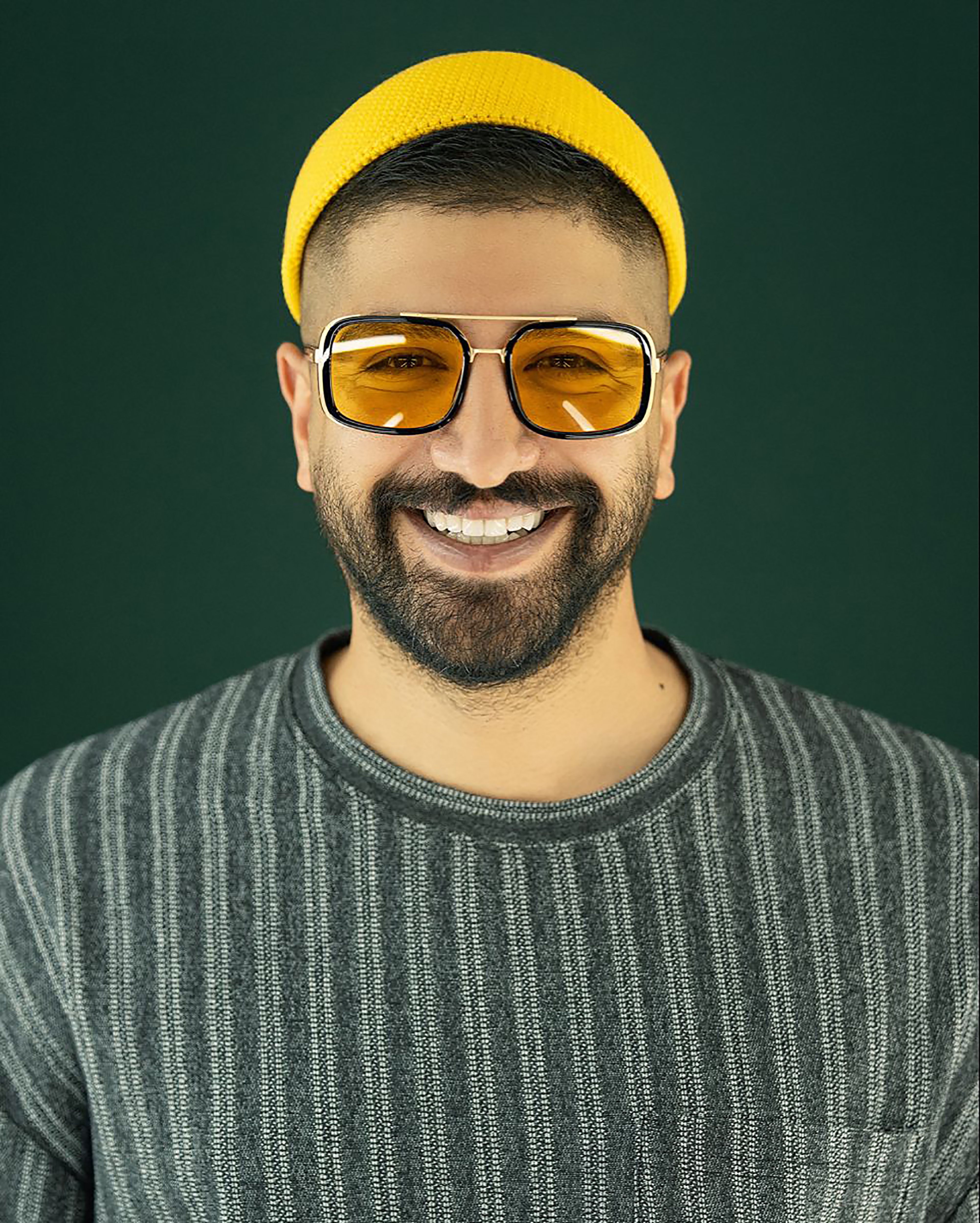 Portrait of a bearded man wearing a yellow beanie and sunglasses with a joyful smile.