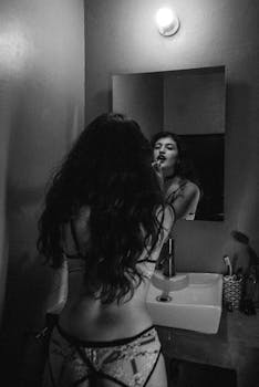 A woman with long hair applies lipstick while looking in the mirror in a grayscale image.