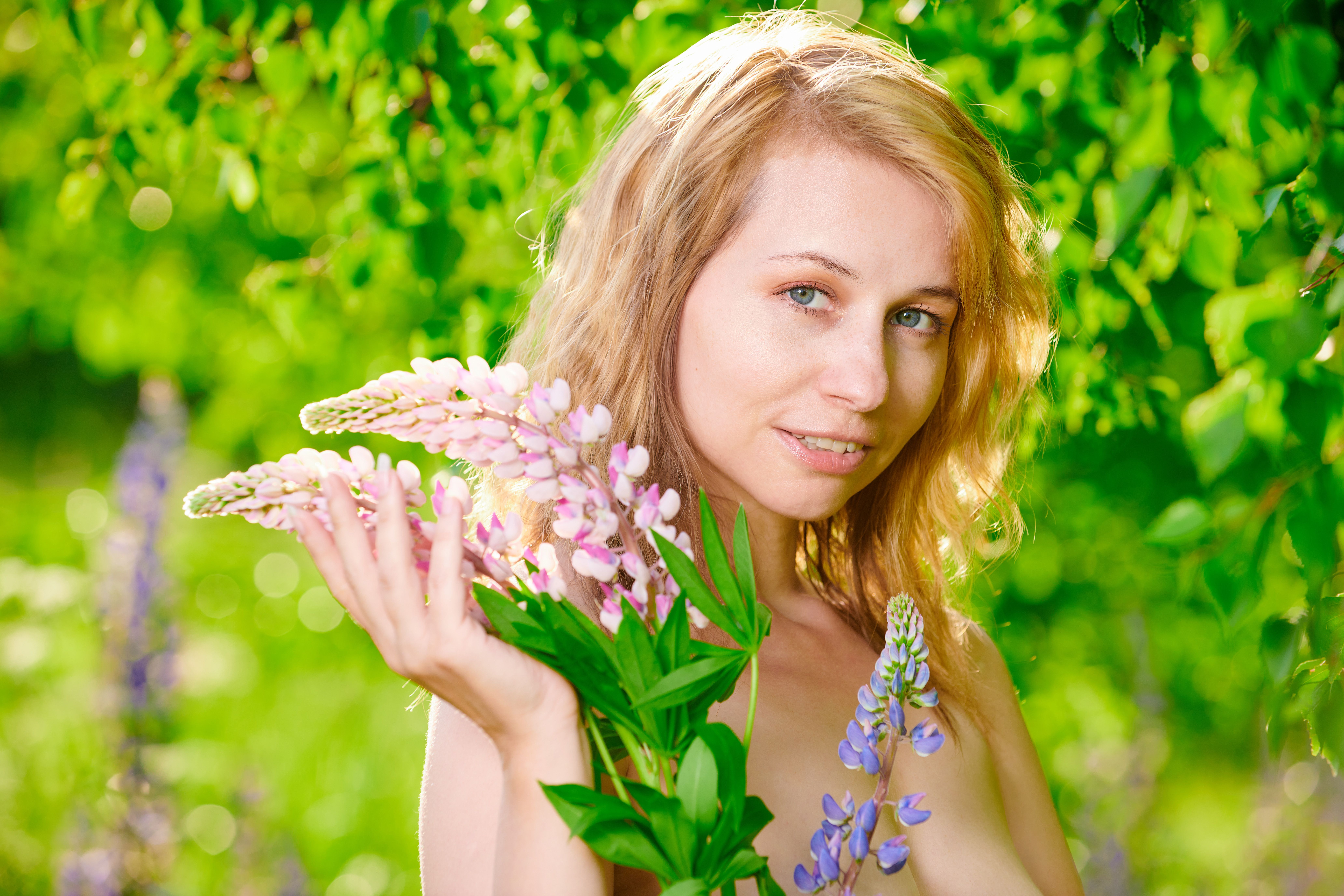 Caucasian woman holding lupine flowers amidst lush greenery on a sunny day.