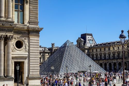 Free Tourists gather around the iconic glass pyramid at the Louvre Museum in Paris under clear blue skies. Stock Photo